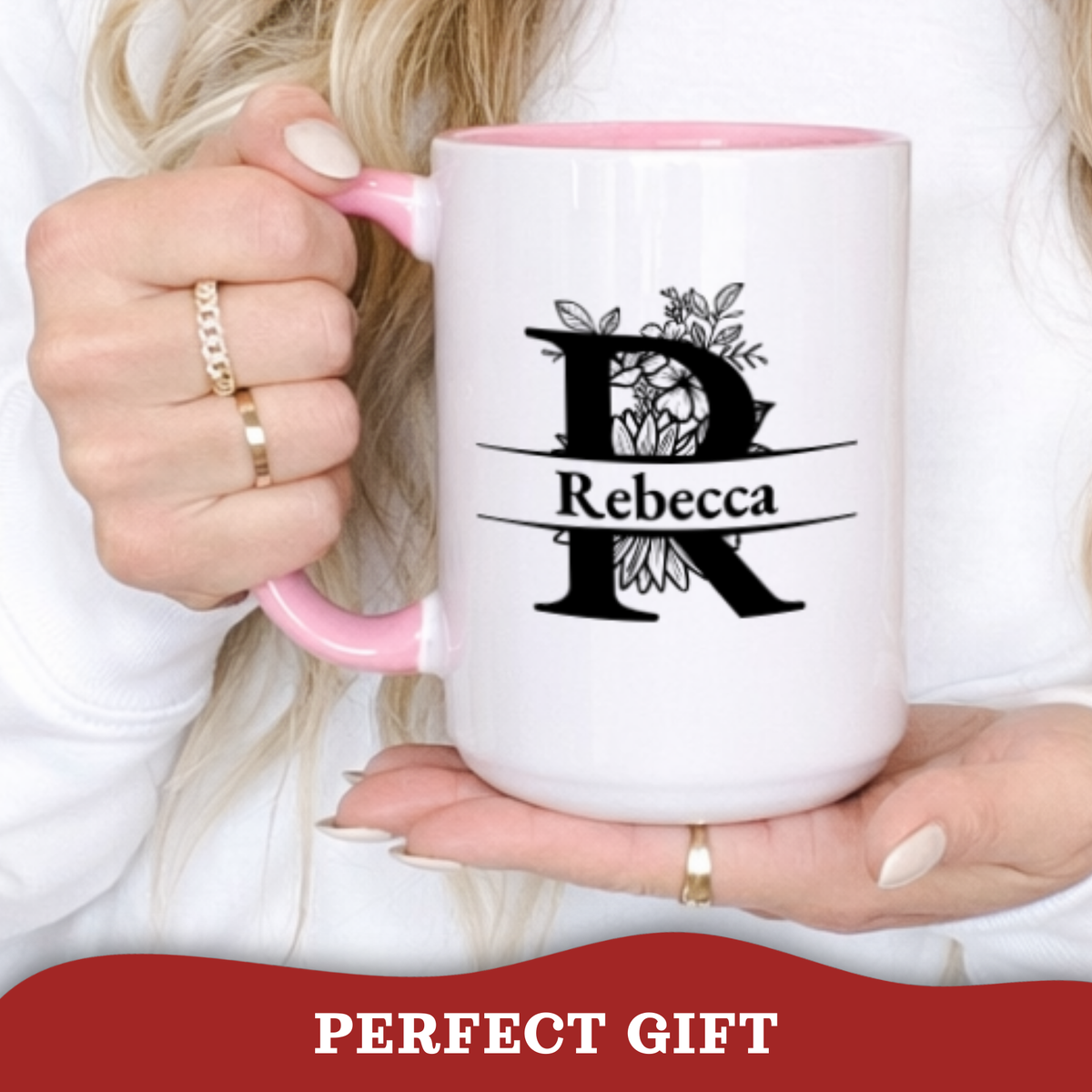 A person holds a white ceramic mug with a pink handle and interior. The mug has a black printed monogram design with the name 'Rebecca' across the center.