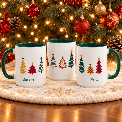 Three personalized Christmas 15oz mugs with green handles and interiors are displayed on a cream knit placemat in front of a lit Christmas tree. The design features colorful tree illustrations around the mug, and the names 'Susan' and 'Eric'.