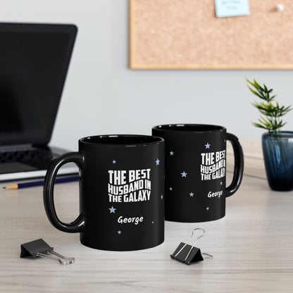 A pair of two black 11oz mugs with small light blue stars and white text 'Best Husband in the Galaxy' with a personalize name below. Showing both sides of the mug. Placed on a wooden surface with paperclips, a pencil and a laptop in the background