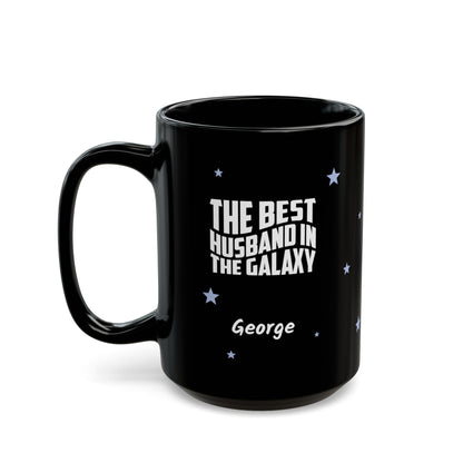 Black 15oz mug with small light blue stars and white text 'Best Husband in the Galaxy' with a personalize name below. Left handle view. Mug is placed on a white background