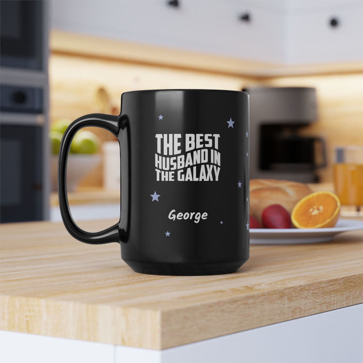 A black 15oz ceramic mug with white text reading 'THE BEST HUSBAND IN THE GALAXY' and the name 'George' printed on it, sitting on a wooden countertop in a blurred kitchen setting.
