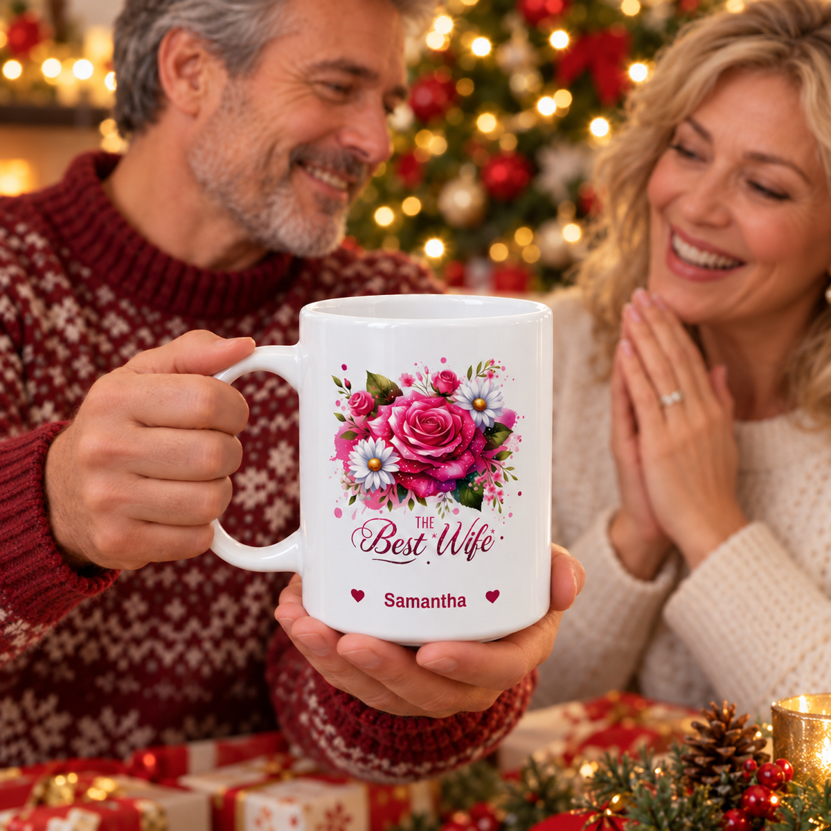 A white ceramic 11oz mug with a pink rose and daisy floral design and the text 'The Best Wife' and 'Samantha' is held by a man in a red sweater, with a smiling woman in a cream sweater and a Christmas tree in the background.