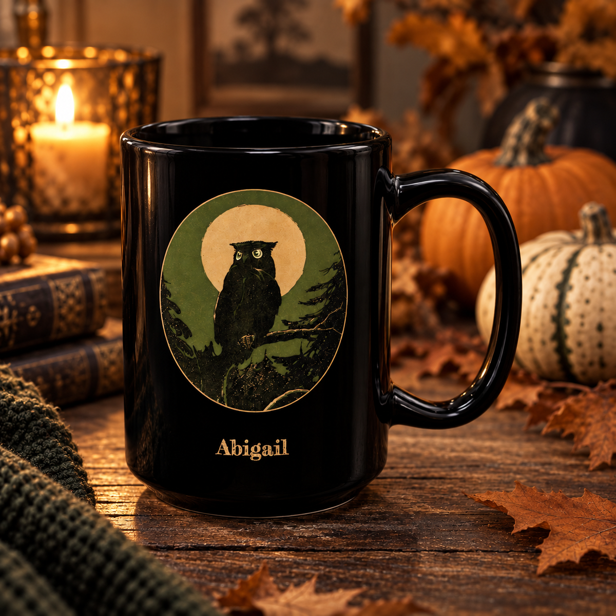 A black ceramic 15oz mug with a printed owl illustration in a circular green and gold color design, with the name 'Abigail' below it, set on a rustic wooden table with autumn decor in the background.