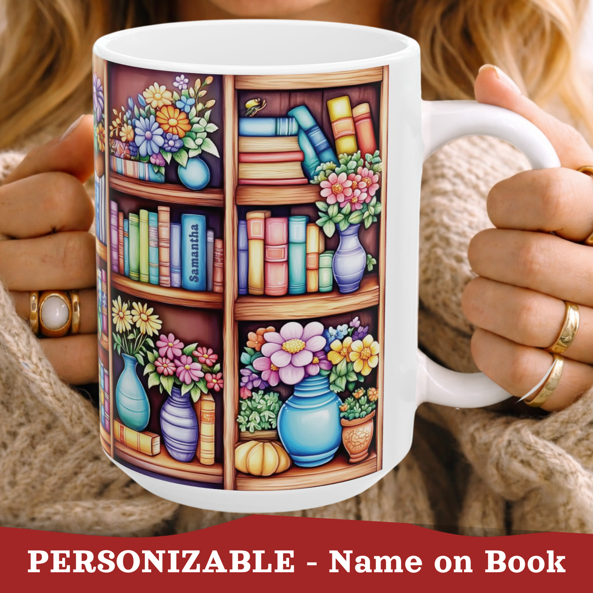 A white ceramic mug with a colorful printed illustration of bookshelves, flowers, and books, held by a person wearing a beige knit sweater. The text at the bottom reads 'PERSONIZABLE - Name on Book'.