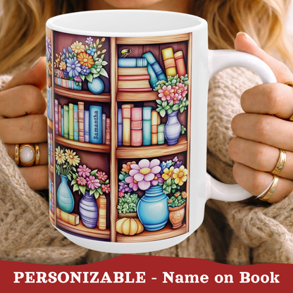 A white ceramic mug with a colorful printed illustration of bookshelves, flowers, and books, held by a person wearing a beige knit sweater. The text at the bottom reads 'PERSONIZABLE - Name on Book'.