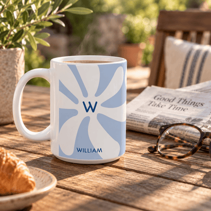 A white ceramic 15oz mug with a light blue sunburst design featuring the initial 'W' and the name 'WILLIAM' printed on it, sitting on a wooden table in a cozy outdoor setting with a croissant, newspaper, and eyeglasses nearby. Left handle view.