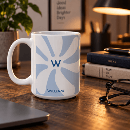 A white ceramic mug with a light blue and white abstract sunburst design, personalized with a blue monogram 'W' and the name 'WILLIAM', sitting on a wooden desk in a warm, cozy office setting.