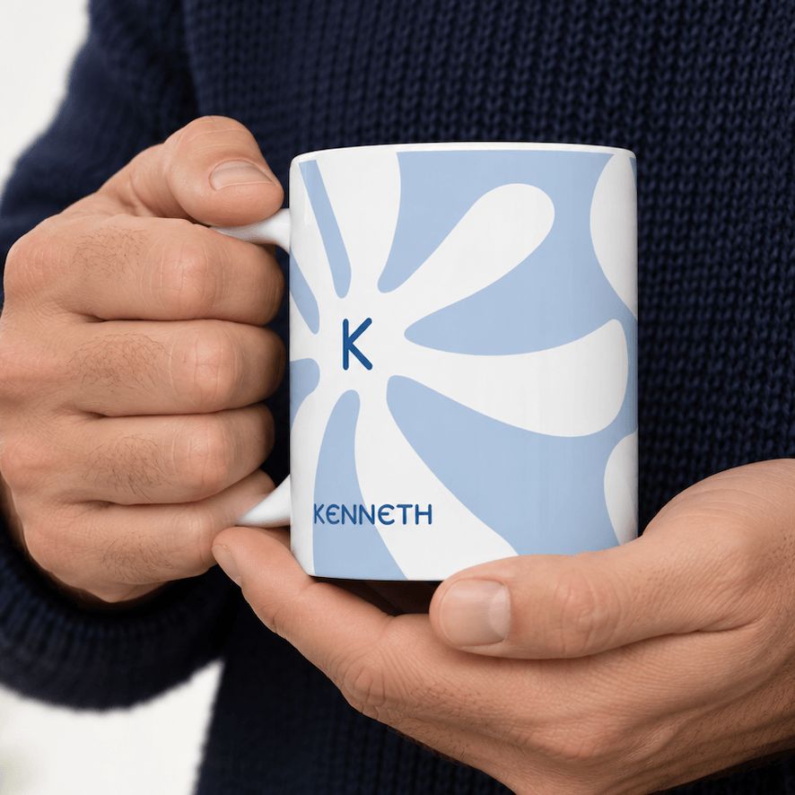 A person in a navy knit sweater holds a white ceramic mug with a light blue abstract floral pattern, a blue initial 'K', and the name 'KENNETH' printed on it.