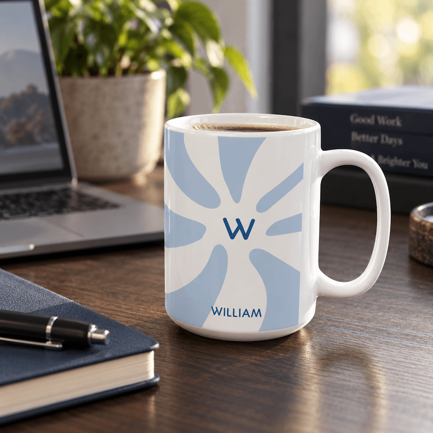 A white ceramic coffee mug with a light blue sunburst pattern featuring the initial 'W' and the name 'WILLIAM' printed in blue, sitting on a wooden desk next to a laptop, notebook, and plant.
