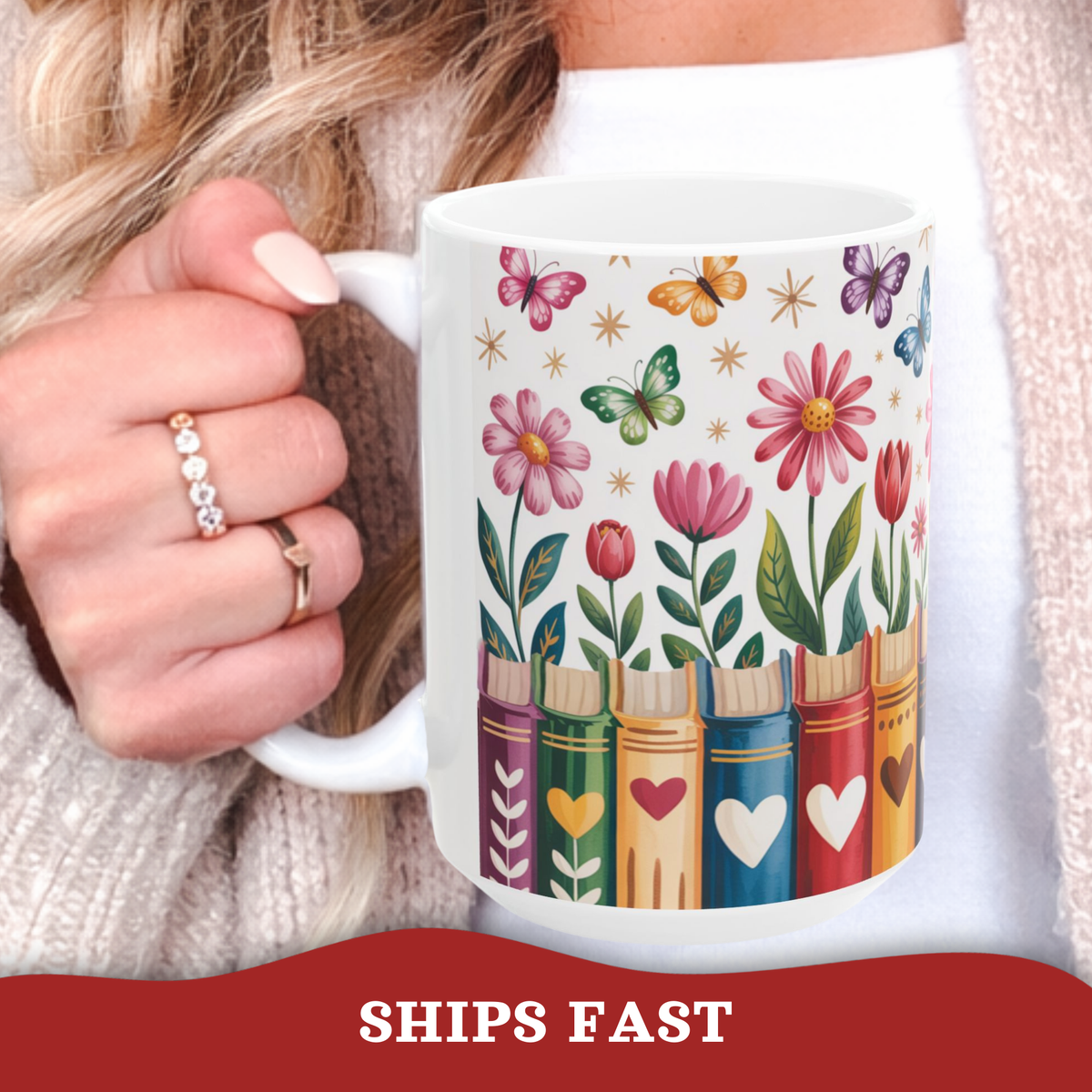 A white ceramic mug with a colorful book-themed floral print, held by a person wearing a beige knit sweater and gold rings. A red banner at the bottom reads 'SHIPS FAST'.