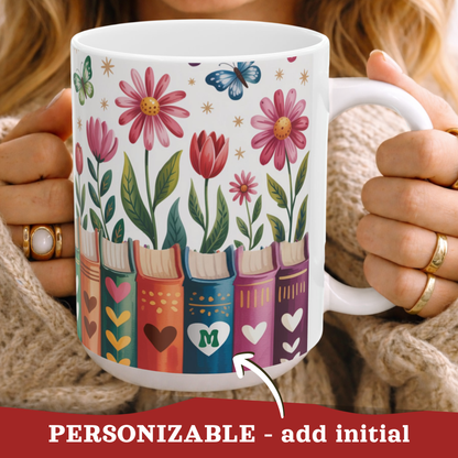 A white ceramic mug with a colorful floral and book illustration, held by a person wearing a beige knit sweater. The mug features pink daisies, red tulips, green leaves, butterflies, and books decorated with hearts and initials. A personalized initial in a heart, on a book spine.