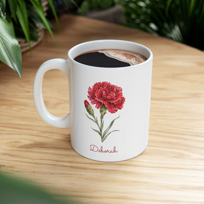 Carnation birth flower personalized ceramic mug, 11oz, on a wooden table with green plant leaves in background.
