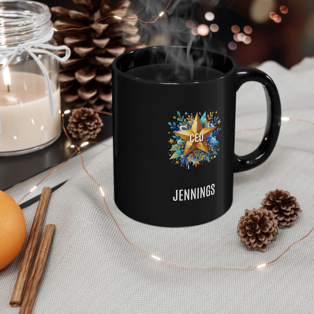 A black 11oz ceramic mug with a custom printed design featuring a gold star, blue floral accents, the text 'CEO' in the center, and personalized name 'JENNINGS' printed below in white. Right handle view. The mug is placed on a linen napkin, surrounded by cone pines, 2 cinnamon sticks, a burning candle and tiny christmas lights.