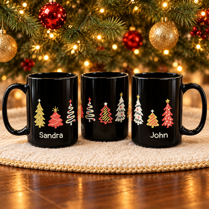 Three black ceramic mugs with personalized names, 'Sandra' and 'John', and colorful Christmas tree designs around the mugs, displayed on a textured cream placemat in front of a glowing Christmas tree.