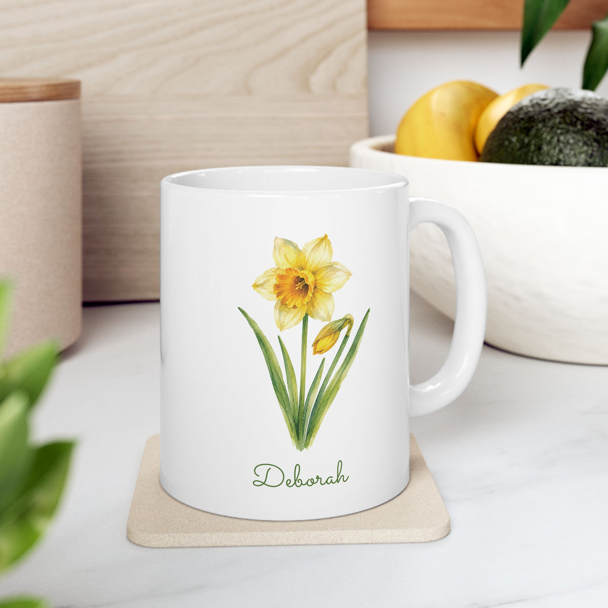 Daffodil birth flower personalized ceramic mug, 11oz, right side view, on a wooden coaster with fruit bowl in background.