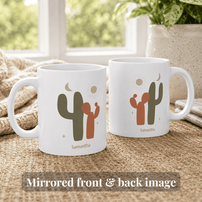 Two white ceramic 11oz mugs with a mirrored desert cactus design, shown on a woven table runner in front of a window. The mugs feature a green and dark orange cactus, a sun, a moon, and the name 'Samantha' printed below.