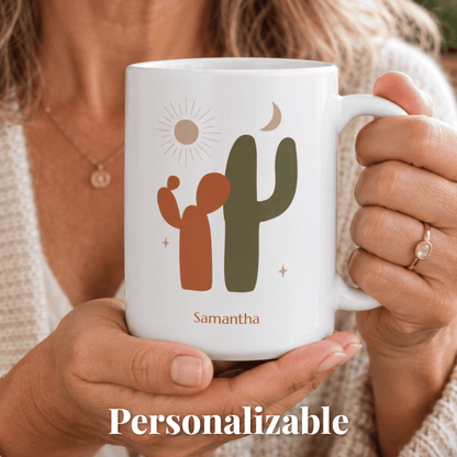 A person holds a white ceramic 15oz mug with a minimalist desert design featuring a cactus, sun, moon, and the name 'Samantha' in brown text. The mug is shown in a close-up lifestyle shot with a 'Personalizable' text overlay at the bottom.