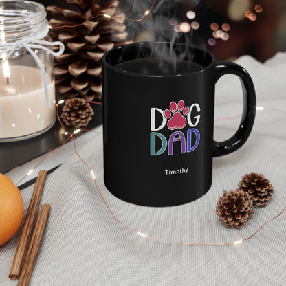 A black ceramic 11oz mug with a 'Dog Dad Timothy' graphic, styled on a light fabric surface with pinecones, a candle, cinnamon sticks, and fairy lights.