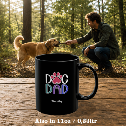 A black ceramic 11oz mug with a colorful 'Dog Dad' paw print graphic and the name 'Timothy' printed on it, shown in the foreground on a wooden surface. The background is a forest scene with a man crouching down to play with a golden retriever dog.
