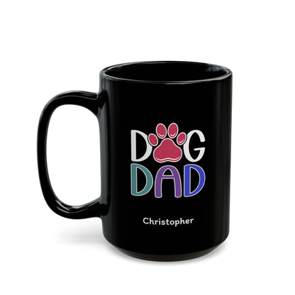 A glossy black 15oz ceramic mug featuring a colorful graphic that reads 'DOG DAD' with a pink paw print and the name 'Christopher' printed below in white, on a plain white background. Left handle view.