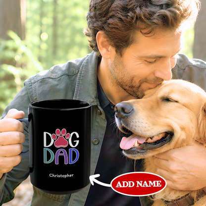 A man in a gray denim shirt holds a black ceramic mug printed with 'DOG DAD' in colorful letters and a pink paw print, while kissing a golden retriever dog outdoors. The image includes a red 'ADD NAME' oval overlay pointing to the mug.