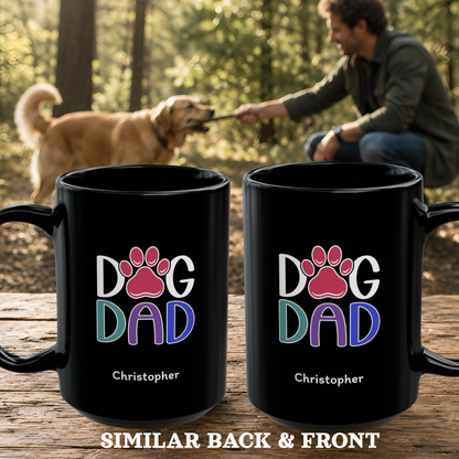 Two black mugs with a colorful 'DOG DAD' paw print design and the name 'Christopher' printed on them, displayed on a wooden surface with a blurred background of a man playing with a dog in a forest.