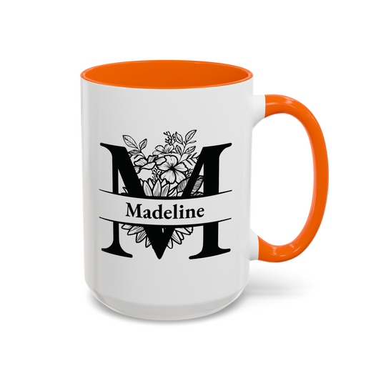 A white ceramic mug with a bright orange handle and interior, featuring a black graphic of a large letter M decorated with floral line art and the name 'Madeline' on a banner.