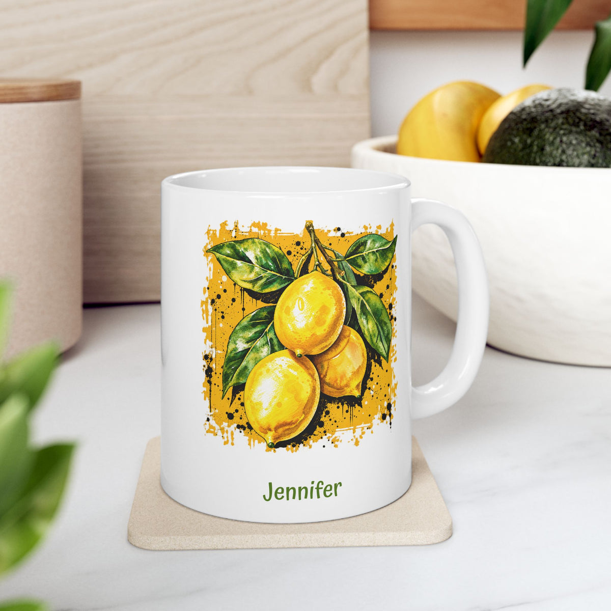 A white ceramic 11oz mug with a printed illustration of three yellow lemons with green leaves on a darker yellow textured background and a name below, set on a wooden countertop in a blurred kitchen environment.