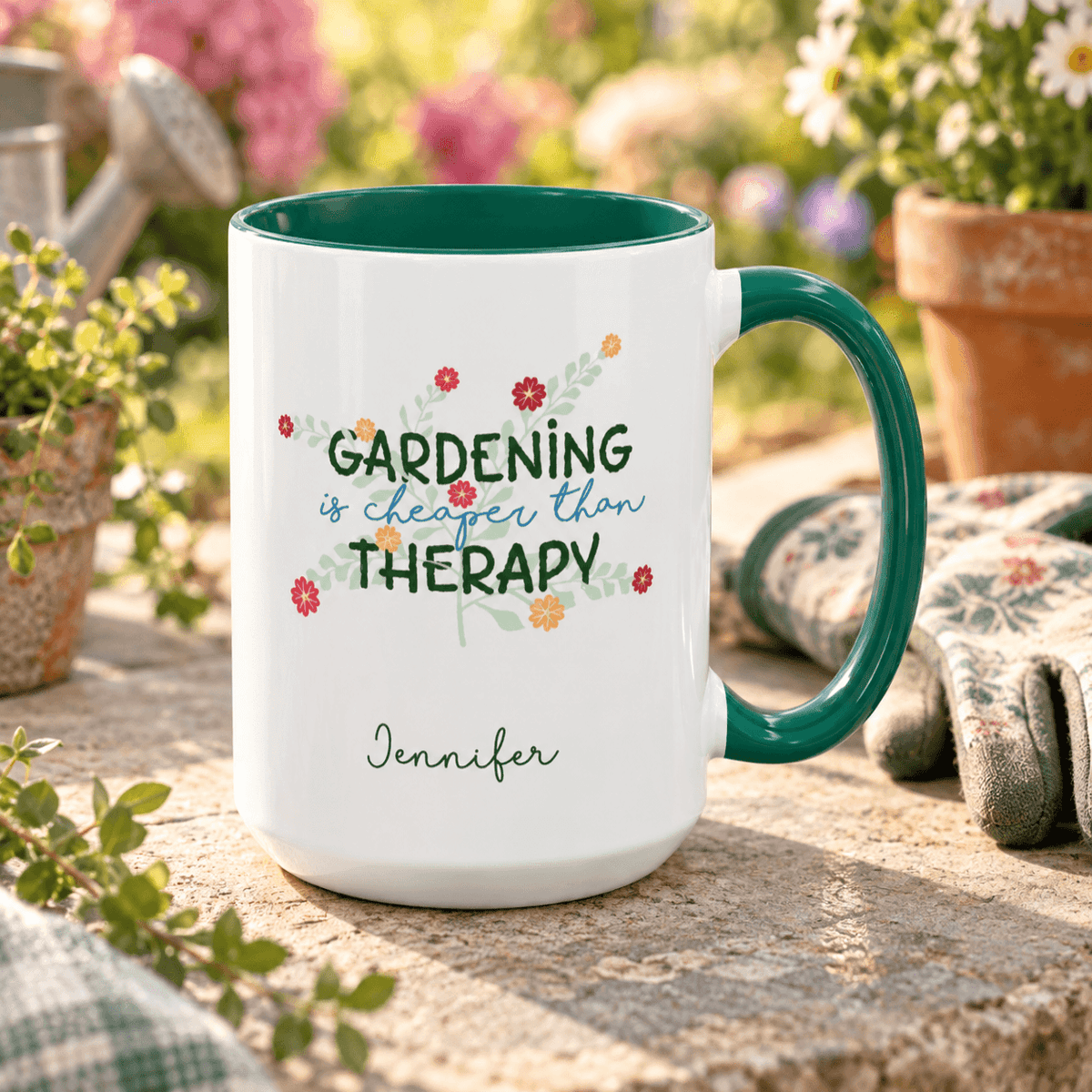 A white ceramic mug with a dark green handle and interior, printed with the phrase 'GARDENING is cheaper than THERAPY' in green and blue script with small floral accents, and the name 'Jennifer' below. The mug is photographed outdoors on a stone surface with gardening gloves, flowers, and a watering can in a blurred garden background.