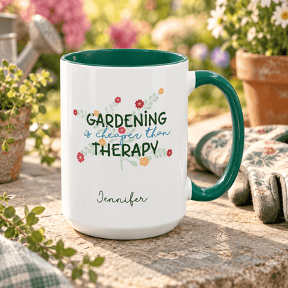 A white ceramic mug with a dark green handle and interior, printed with the phrase 'GARDENING is cheaper than THERAPY' in green and blue script with small floral accents, and the name 'Jennifer' below. The mug is photographed outdoors on a stone surface with gardening gloves, flowers, and a watering can in a blurred garden background.