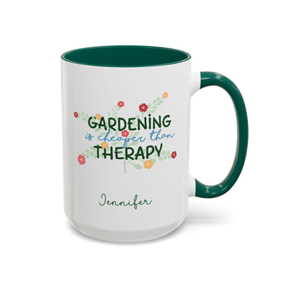 A white ceramic 15oz mug with a dark green handle and interior, featuring the printed phrase 'GARDENING is cheaper than THERAPY' in green and blue script with small colorful flowers, and the name 'Jennifer' in green cursive below. Right handle view.