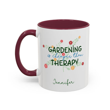 A white ceramic 11oz mug with a maroon colored handle and interior, featuring the printed phrase 'GARDENING is cheaper than THERAPY' in green and blue script with small colorful flowers and the name 'Jennifer' below. Left handle view.