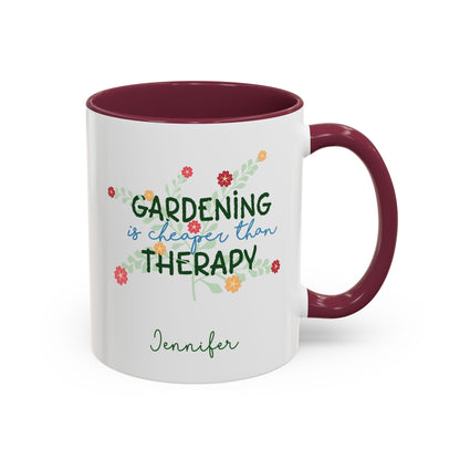A white ceramic 11oz mug with a maroon colored handle and interior, featuring the printed phrase 'GARDENING is cheaper than THERAPY' in green and blue script with small colorful flowers, and the name 'Jennifer' in green cursive below. Right handle view.