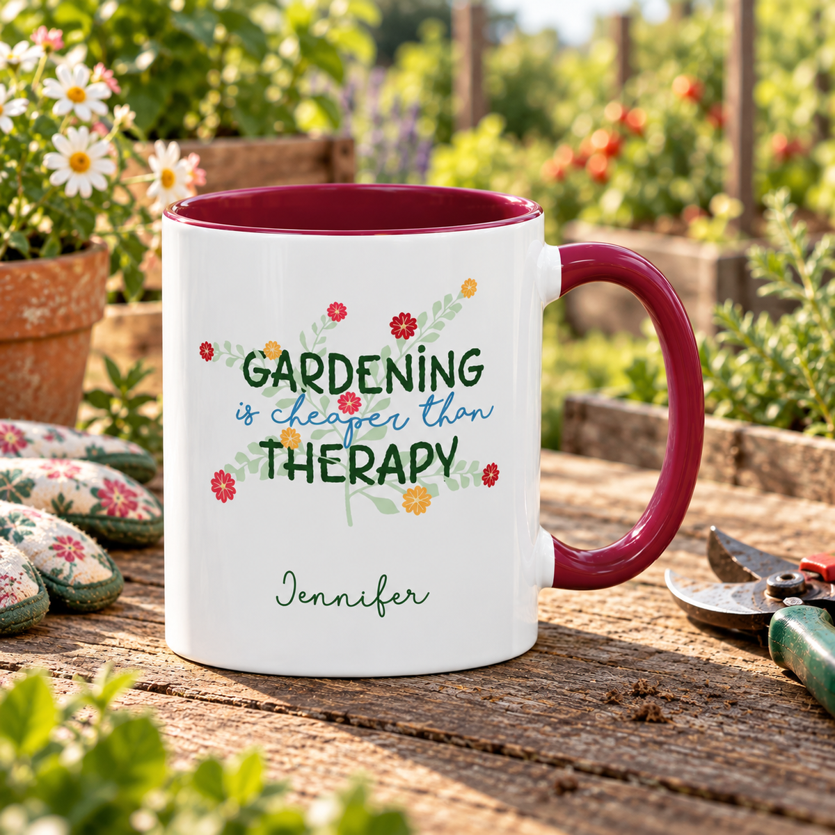 A white ceramic mug with a maroon colored handle and interior, printed with the phrase 'GARDENING is cheaper than THERAPY' in green and blue lettering with small floral accents, and the name 'Jennifer' below. The mug is placed on a wooden table in a garden setting with flowers, gardening gloves, and pruning shears visible.