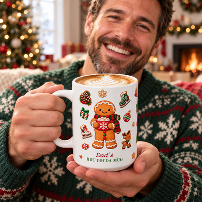 A smiling man in a green holiday sweater holds a white ceramic 15oz mug printed with gingerbread-themed illustrations and the text 'Dad's HOT COCOA MUG' in a cozy indoor setting with a blurred Christmas tree and fireplace in the background.