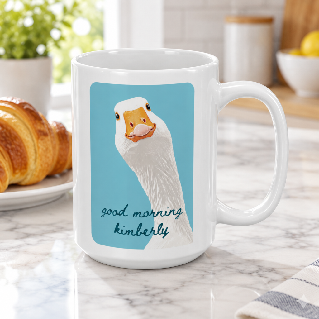 A white ceramic 15oz mug with a printed illustration of a white goose on a light blue background and the text 'good morning kimberly' in dark teal script. The mug is placed on a marble countertop in a kitchen setting with a croissant and other breakfast items blurred in the background.