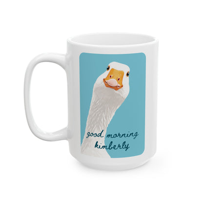 A white ceramic 15oz mug featuring a printed graphic of a white goose with an orange beak against a light blue square, with the customizable text 'good morning kimberly' in dark blue script below. Left handle view.