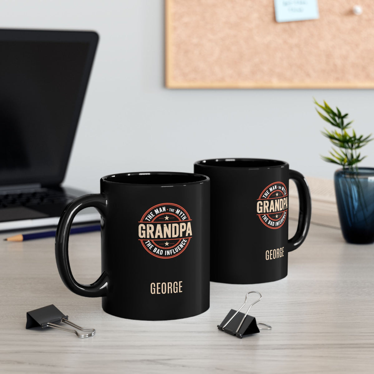 A pair of two black 11oz mugs with 'Grandpa: The Man, The Myth, the Bad Influence' text placed in a circle, and personalized name below. Showing both sides of the mug. Placed on a wooden surface with paperclips, a pencil and a laptop in the background