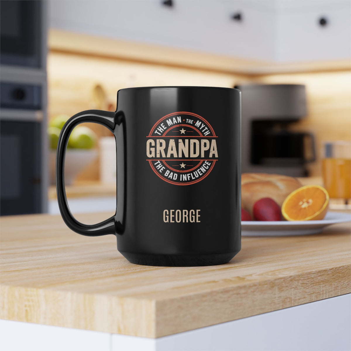 A black 15oz ceramic mug with 'Grandpa: The Man, The Myth, the Bad Influence' text placed in a circle, and the name 'George' printed below, sitting on a wooden countertop in a blurred kitchen setting.
