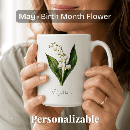 A woman holds a white ceramic mug with lily of the valley birth flower and personalized name at bottom.