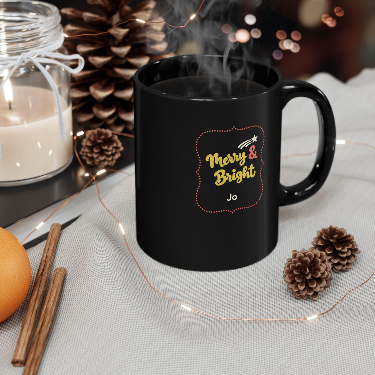 Glossy black 15oz mug with yellow and coral red design with text 'Merry & Bright' in thick yellow script lettering and a personalizable name. Mug is placed on a light grey tablecloth with cinnamon sticks, christmas lights and a candle.