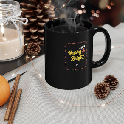 Glossy black 15oz mug with yellow and coral red design with text 'Merry & Bright' in thick yellow script lettering and a personalizable name. Mug is placed on a light grey tablecloth with cinnamon sticks, christmas lights and a candle.