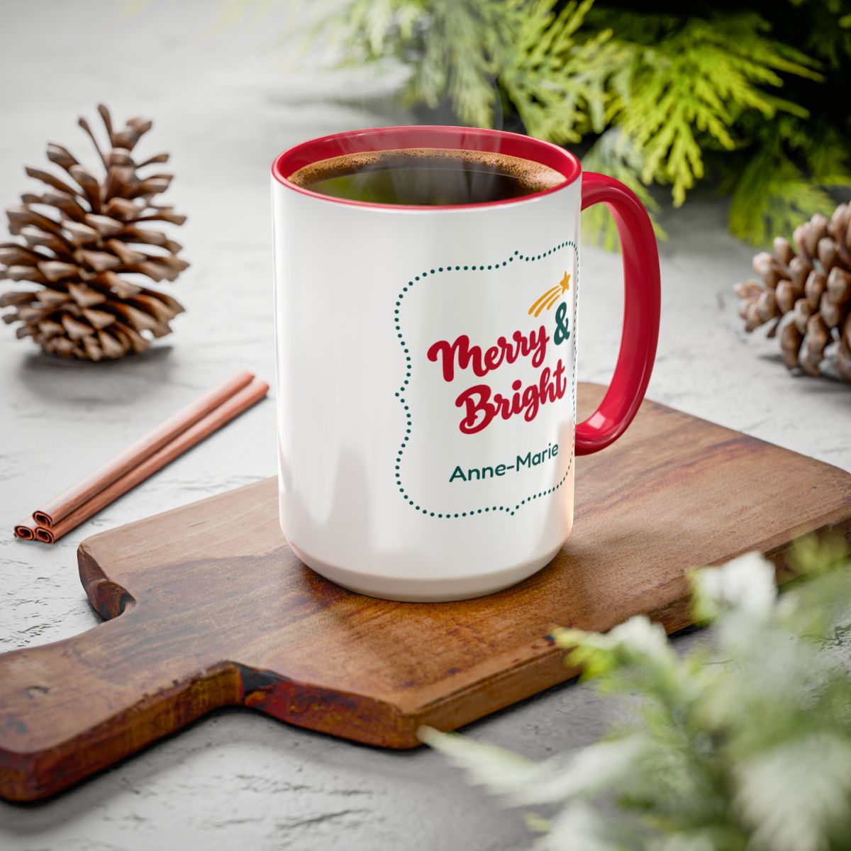 A white ceramic 15oz mug with a red handle and rim, filled with coffee, sitting on a wooden cutting board. The mug features a 'Merry & Bright' holiday graphic in bold red script lettering, and a personalizable name 'Anne-Marie'.