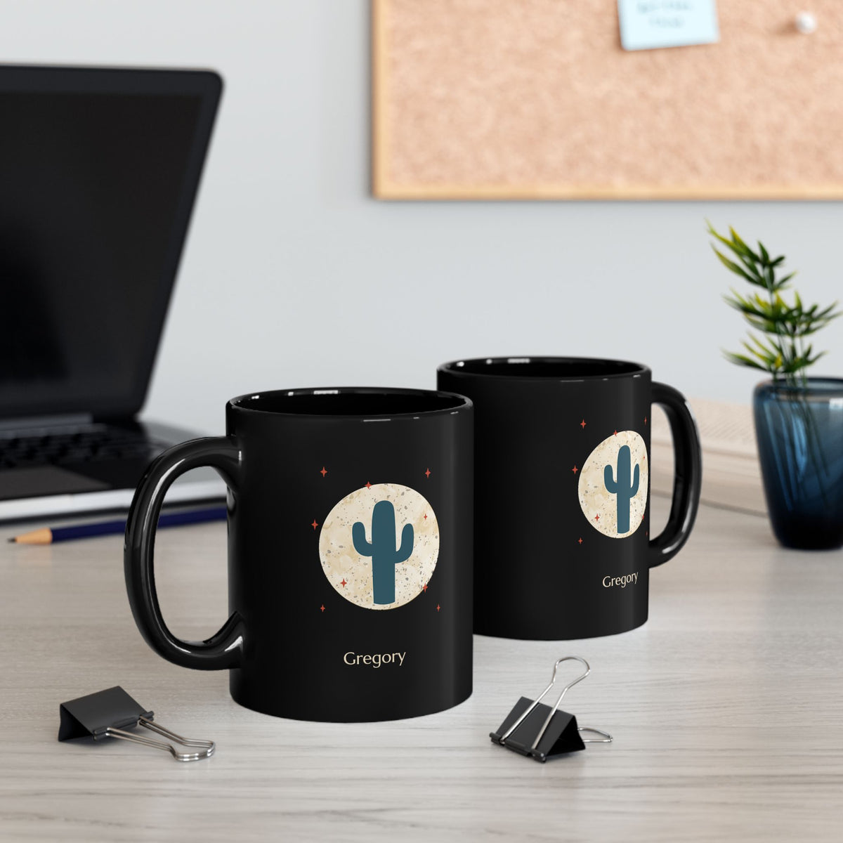 Two black 11oz black ceramic mugs, featuring a printed graphic of a teal cactus inside a beige circle with small red stars, and the name 'Gregory' printed below. The mug is set on a light wooden desk with a blurred laptop and paperclips in the background.