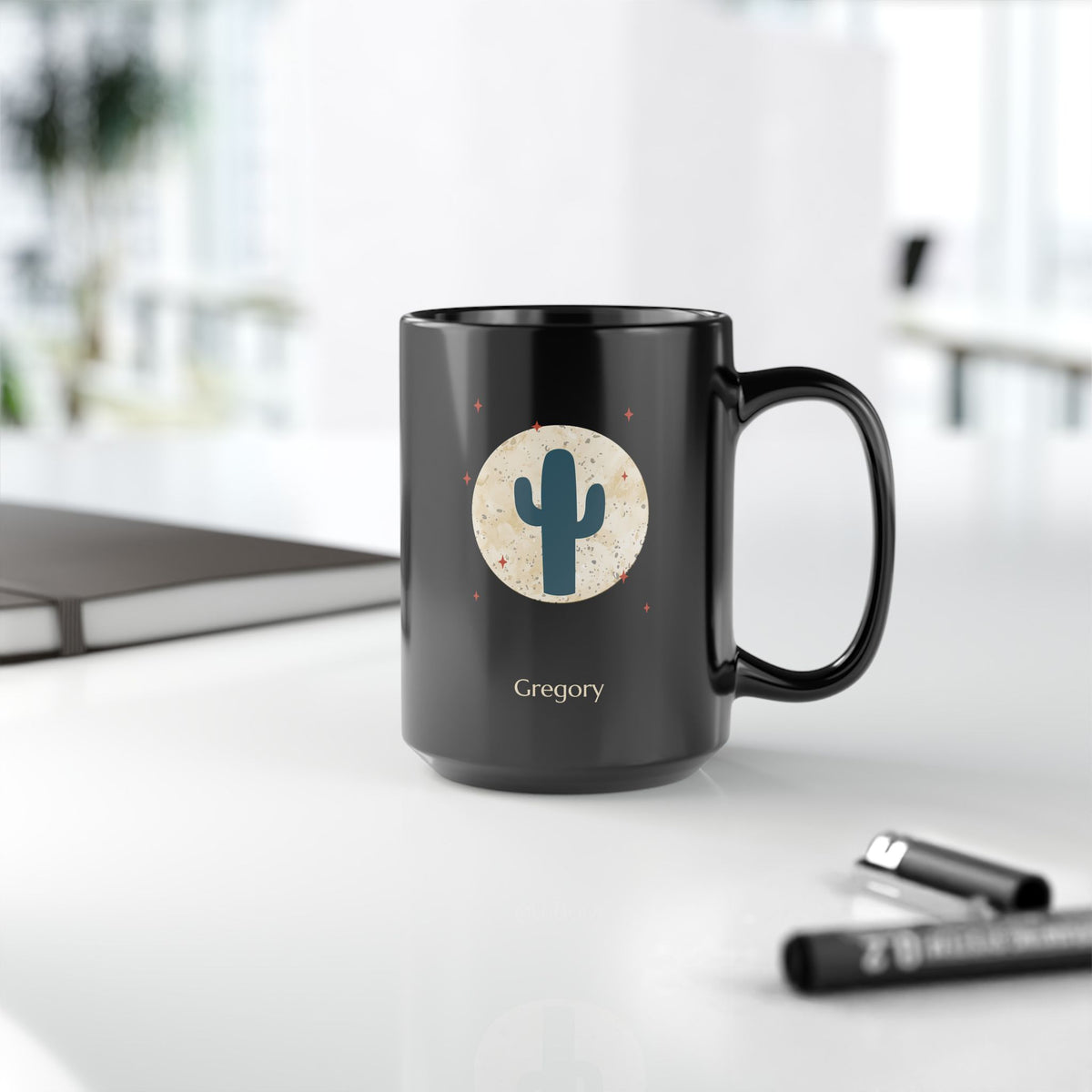 A black 15oz ceramic mug with a handle, featuring a printed graphic of a teal cactus inside a beige circle with small red stars, and the name 'Gregory' printed below. The mug is set on a white desk with a blurred laptop and pens in the background, right handle view.