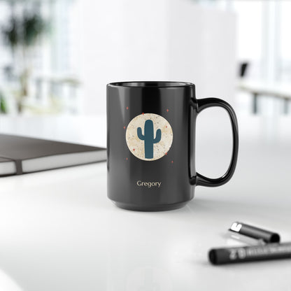 A black 15oz ceramic mug with a handle, featuring a printed graphic of a teal cactus inside a beige circle with small red stars, and the name 'Gregory' printed below. The mug is set on a white desk with a blurred laptop and pens in the background, right handle view.