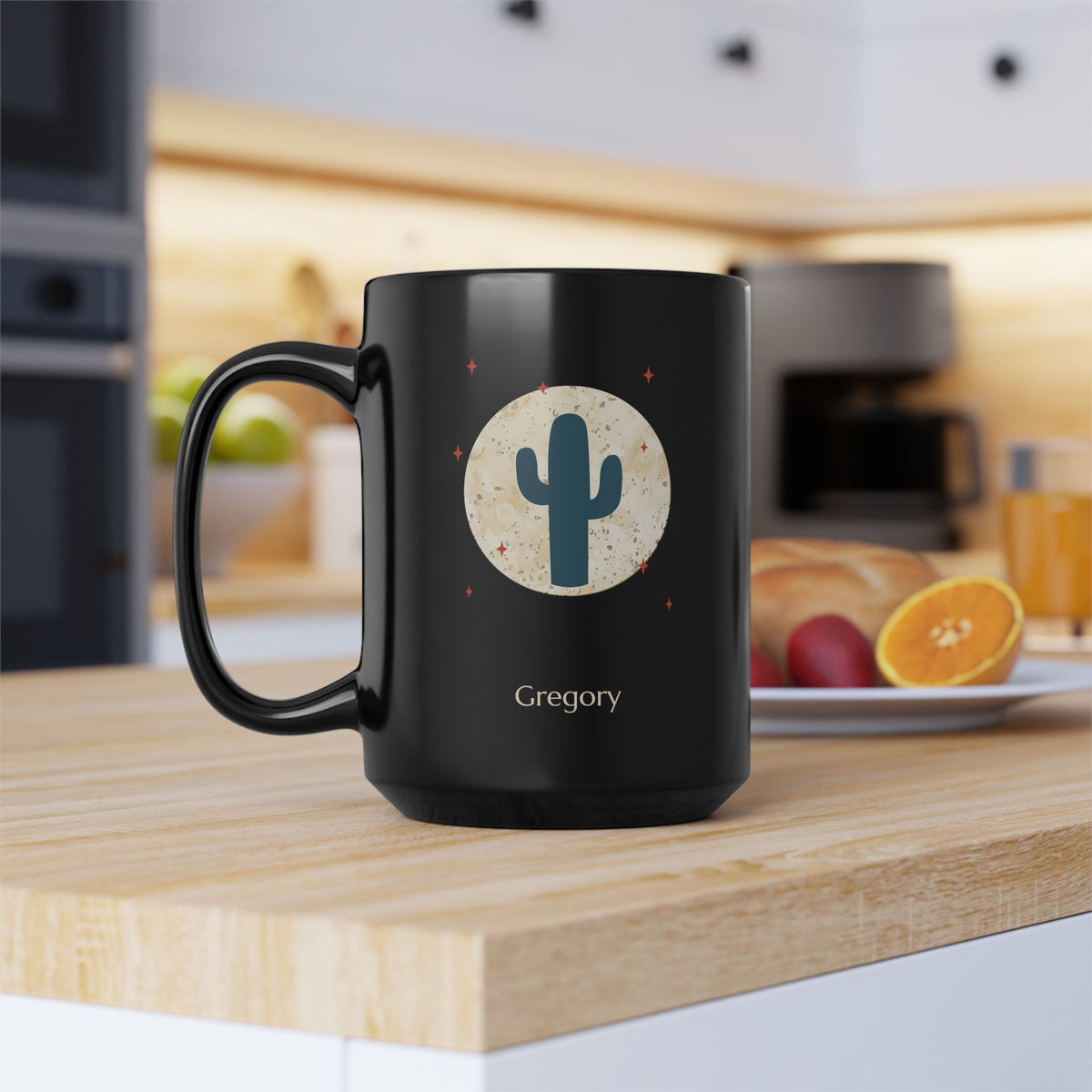 A 15oz glossy black ceramic mug, featuring a printed graphic of a teal cactus inside a beige circle with small red stars, and the name 'Gregory' printed below. Placed on a wooden kitchen table with orange and red fruit on a plate in the background, left handle view.