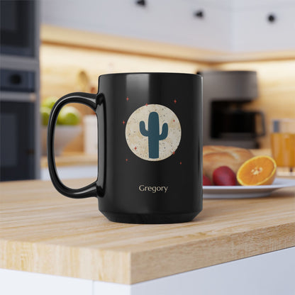 A 15oz glossy black ceramic mug, featuring a printed graphic of a teal cactus inside a beige circle with small red stars, and the name 'Gregory' printed below. Placed on a wooden kitchen table with orange and red fruit on a plate in the background, left handle view.