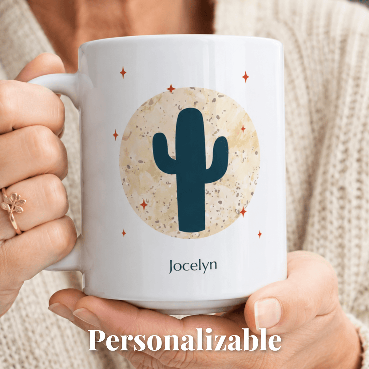 A person holds a white ceramic mug with a desert-themed graphic featuring a dark blue cactus, a beige circular sun, and small orange stars. The name 'Jocelyn' is printed below the design.