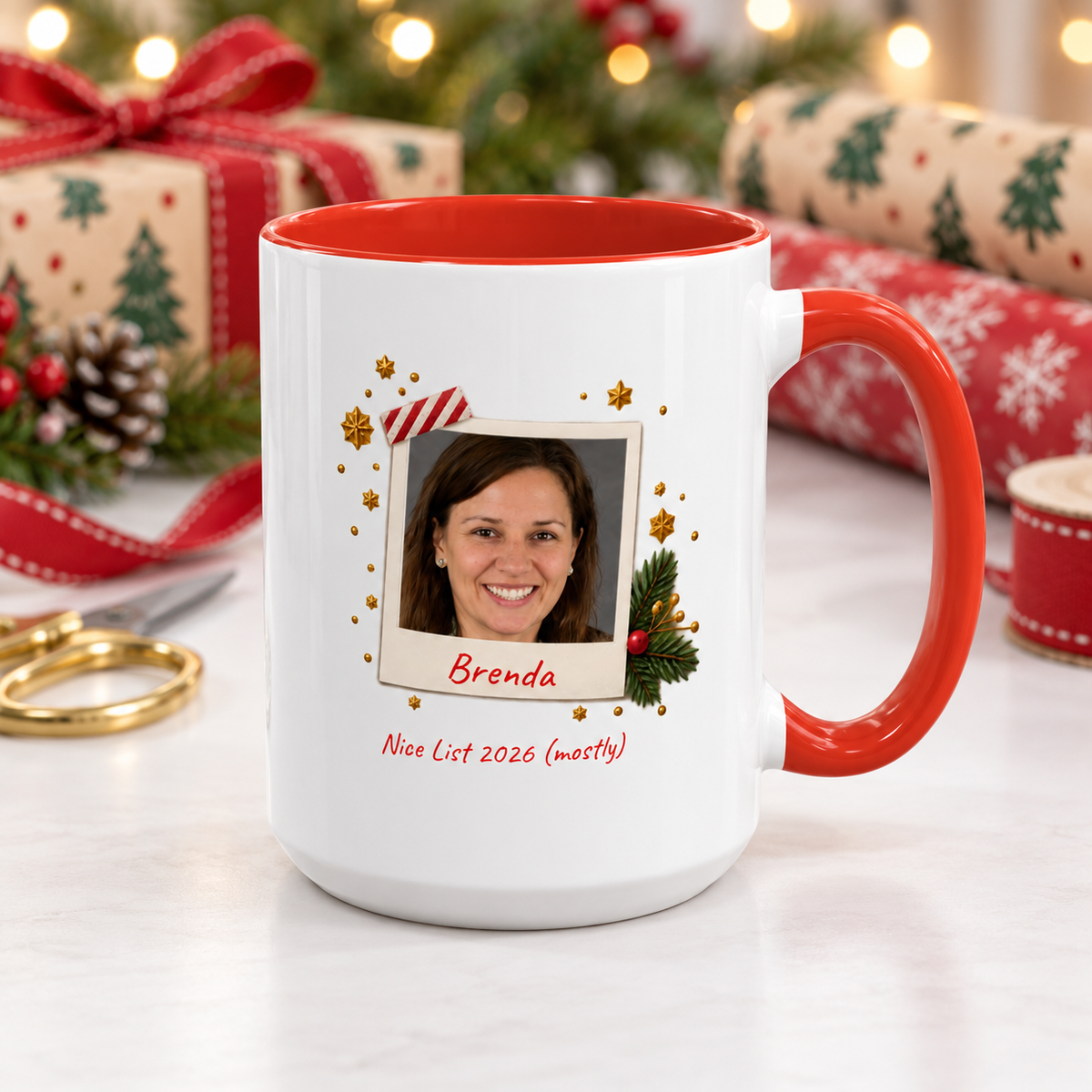 A white ceramic 15oz mug with a red interior and red handle, decorated with a custom polaroid photo frame design, gold stars, and red text reading 'Brenda' and 'Nice List 2026 (mostly)'. The mug is set on a white marble surface with a blurred Christmas background featuring wrapped gifts, a red ribbon, and holiday lights.